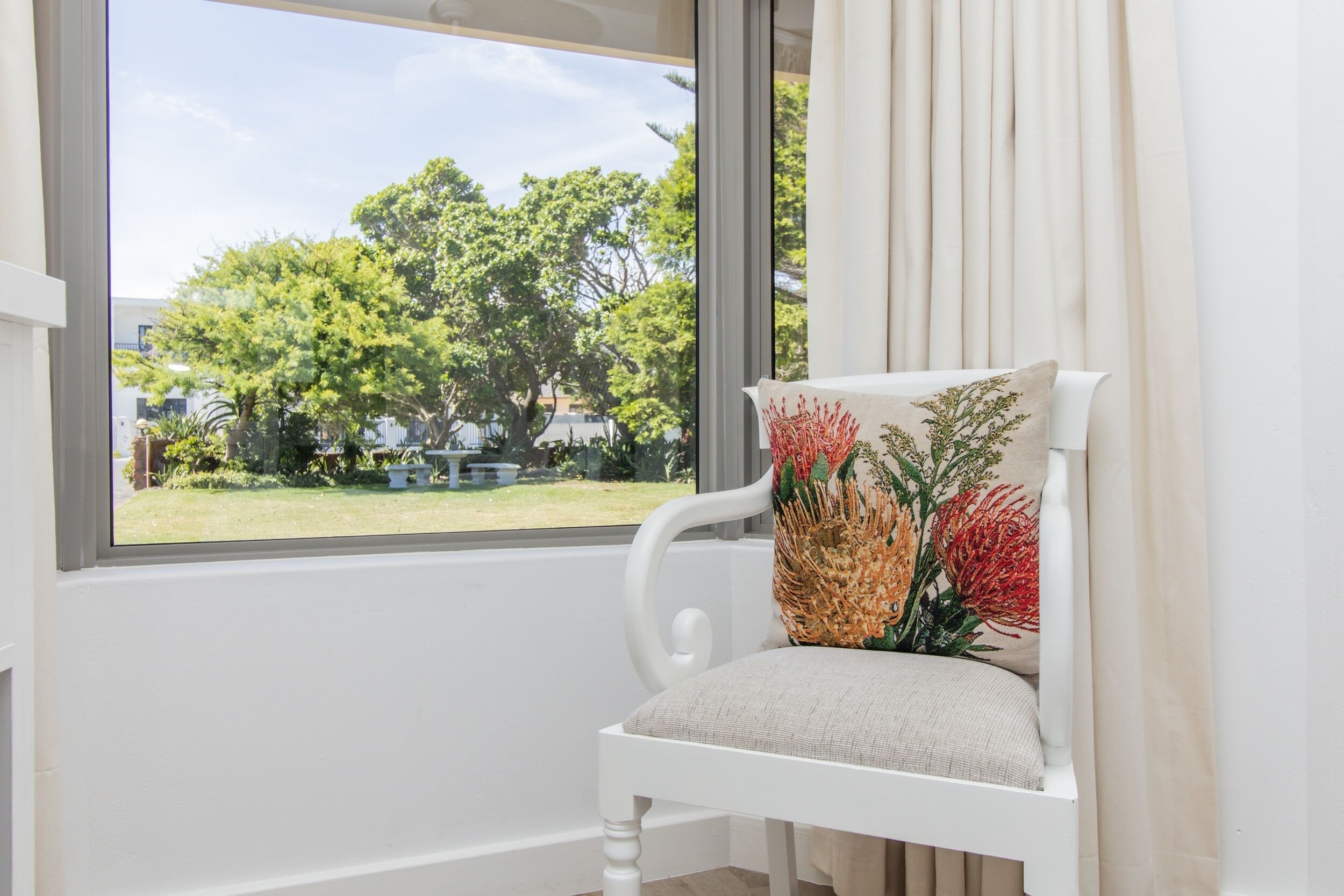 Una silla blanca con un cojín estampado con motivos florales, ubicada junto a una ventana que ofrece vista a un césped verde y árboles.