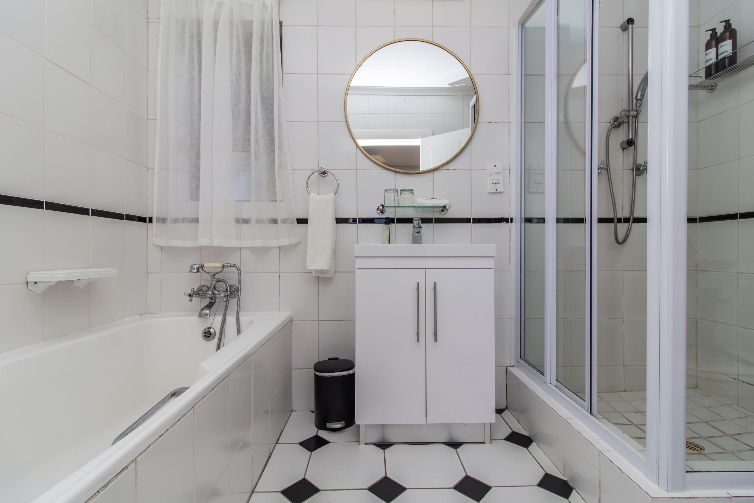 A modern bathroom with a bathtub, a glass-enclosed shower, a white sink with a mirror above it, and a black trash bin.