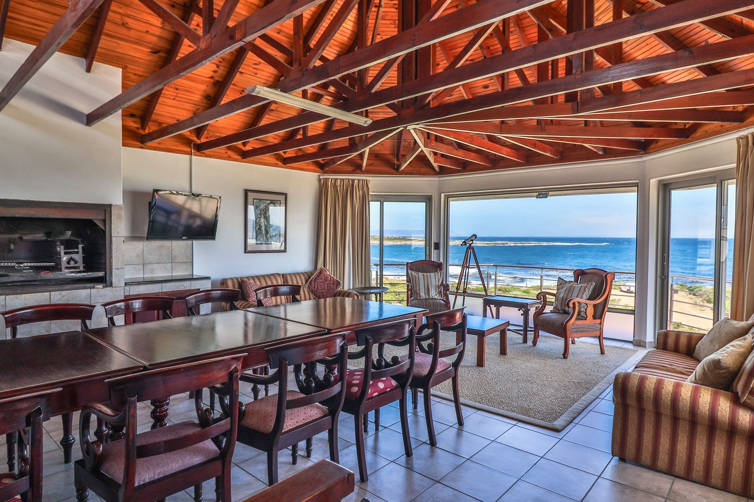 Un comedor con una amplia mesa de madera y sillas, un televisor montado en la pared y vistas al océano a través de grandes ventanales.