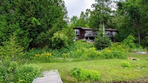 A cabin surrounded by dense forest, a wooden dock extending into a stream, and a clear view of the cabin's exterior. - Four Mile Memories - Three Bedroom Cottage, Sleeps 6 (Kawartha Lakes)