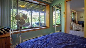Ein Schlafzimmer mit einem Bett, einem Standventilator und Blick ins Freie durch große Fenster.