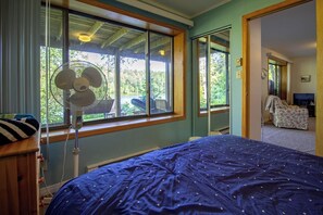 Ein Schlafzimmer mit einem Bett, einem Standventilator und Blick ins Freie durch große Fenster.