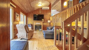 A wooden interior with a fireplace, a TV mounted on the wall, a sofa, and a staircase. - Four Mile Memories - Three Bedroom Cottage, Sleeps 6 (Kawartha Lakes)