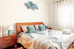 Un letto rifatto con cura, con una coperta a fantasia, un comodino in legno con una lampada e un elemento decorativo appeso al muro.