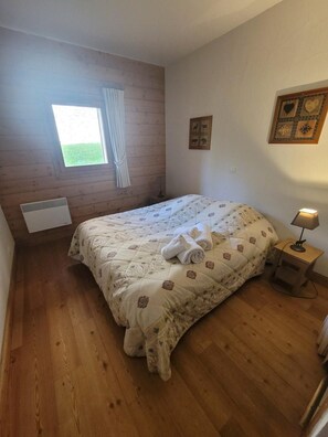 A bedroom with a wooden floor, a bed with a floral duvet, a window with curtains, a wall-mounted picture, and a bedside lamp. - 3rd floor, fitness, swimming pool, sauna, hammam, terrace, parking, tv, 57m², Champagny-en-Vanoise (Champagny-en-Vanoise)