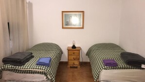 Une chambre avec deux lits simples, chacun recouvert d’une couvre-lit à carreaux et agrémenté d’un oreiller. Une petite table de chevet en bois avec une lampe se trouve entre les deux lits. Un tableau encadré est accroché au mur, au-dessus de la table de chevet.