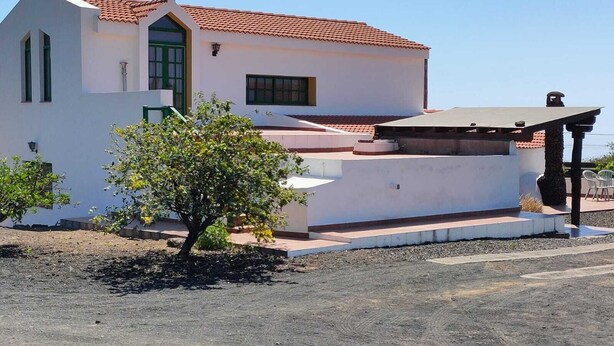 Un bâtiment blanc avec un toit recouvert de tuiles rouges, une terrasse couverte et un petit arbre au premier plan.