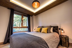 A bedroom with a large bed, a window with curtains, a wooden ceiling, and a nightstand with a lamp.