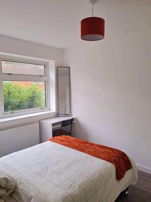 A bedroom with a bed, a desk, a chair, a window, and a red hanging light fixture. - The Happy Place (Cheshunt)