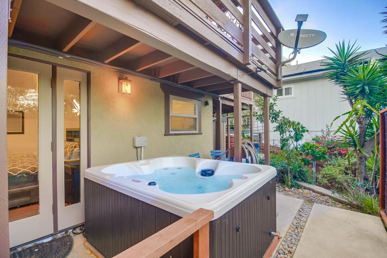 Un jacuzzi en un área exterior cubierta con terraza de madera, antena parabólica y jardín con diversas plantas.