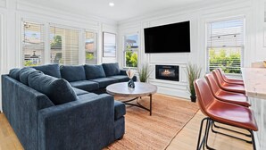 A modern living room with a sectional sofa, a round coffee table, a fireplace, a TV mounted on the wall, and a dining area with bar stools. - San Diego Gem-Charming 4BR Home (San Diego)