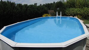 Ein rechteckiger Aufstellpool mit einer Leiter an einer Seite, umgeben von einer Holzterrasse und einem Garten mit verschiedenen Pflanzen und Bäumen.
