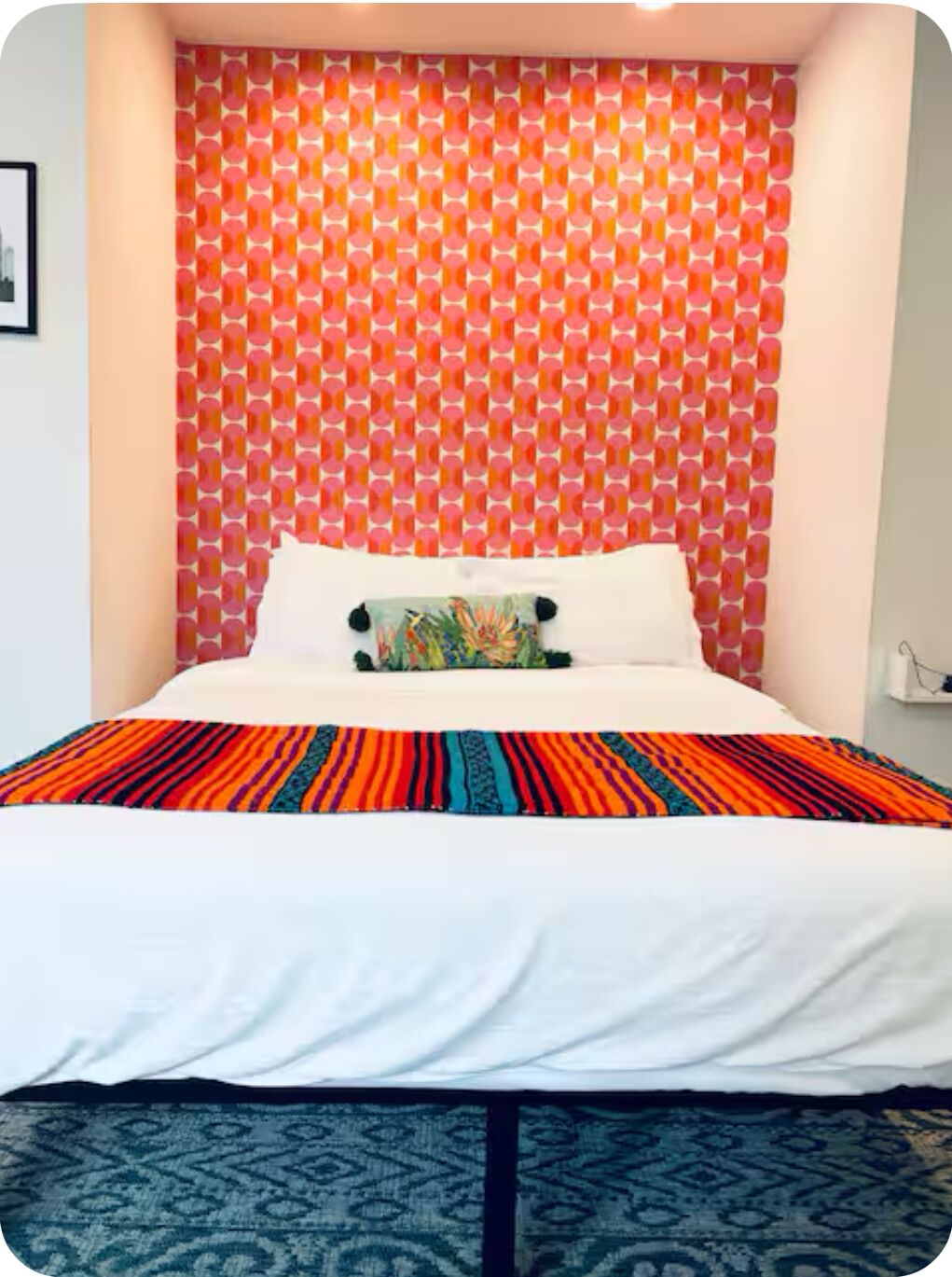 A bed with a colorful striped bedspread, a white pillow, and a patterned pillow. The wall behind the bed has a geometric pattern with circular shapes in shades of pink and orange.