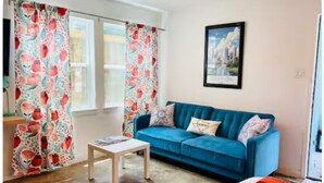 A living room with a blue sofa, a small wooden table, and patterned curtains. - Vintage Chalet Studio 6 Creative Walkable Montrose H-Town Favorite!! (Houston)