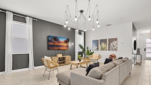 A modern living room with a grey sofa, wooden coffee table, and wicker chairs. There is a TV mounted on the wall and a chandelier above. - 681-Sticks (Kissimmee)