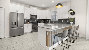 A modern kitchen with white cabinets, stainless steel appliances, a central island with bar stools, and pendant lighting. - 681-Sticks (Kissimmee)