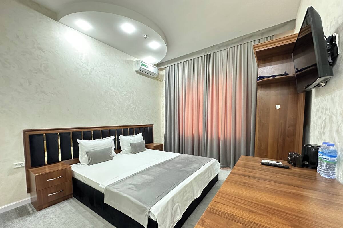 A hotel room with a bed, a wooden desk, a television, and an air conditioning unit.