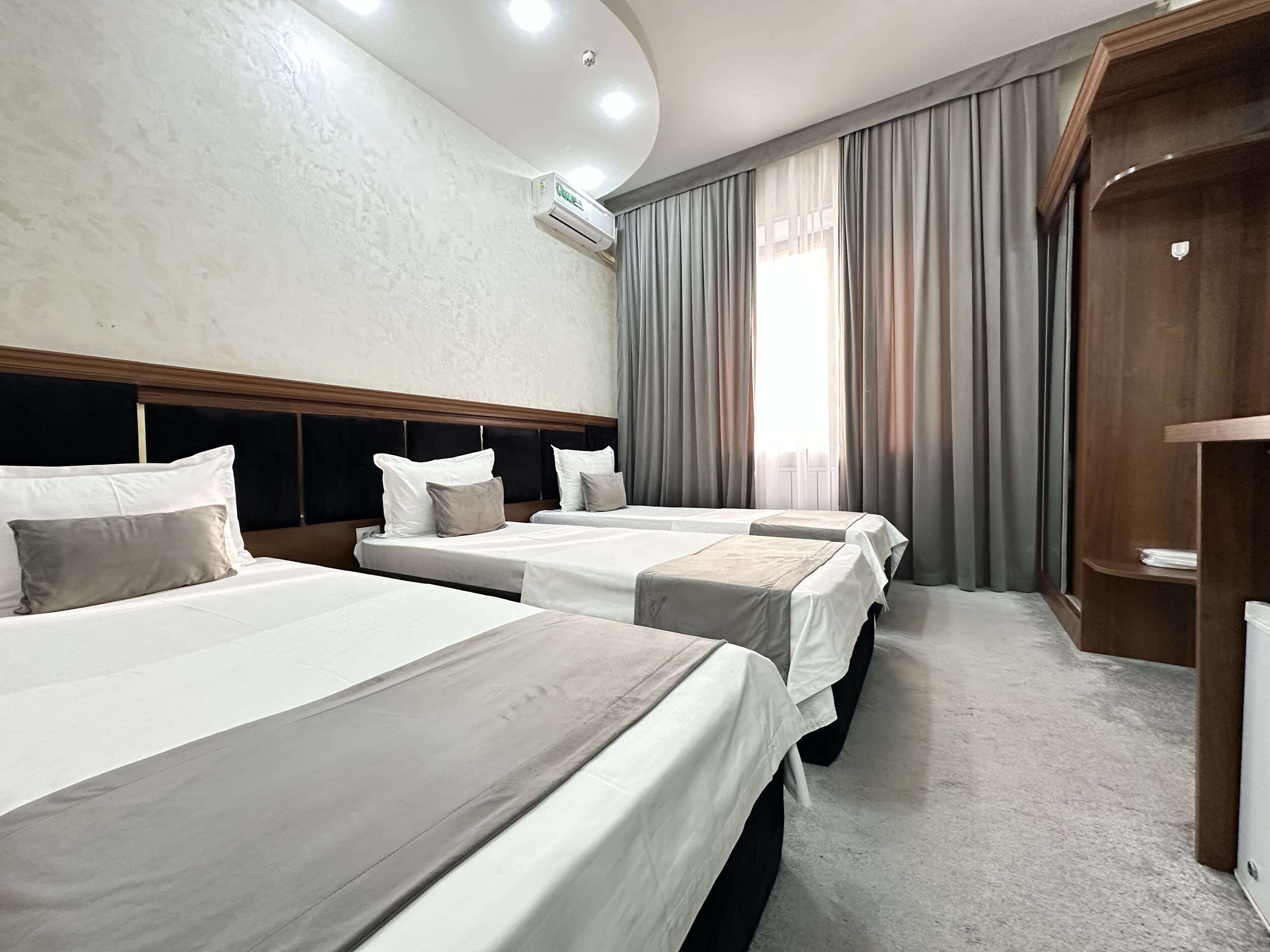 A hotel room with three beds, a wooden headboard, a large window with curtains, and an air conditioning unit on the wall.