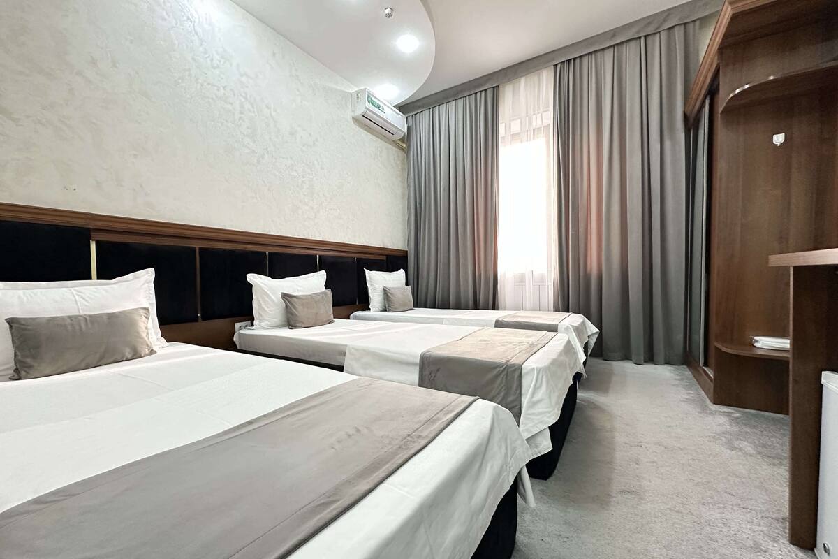 A hotel room with three beds, a wooden headboard, a large window with curtains, and an air conditioning unit on the wall.