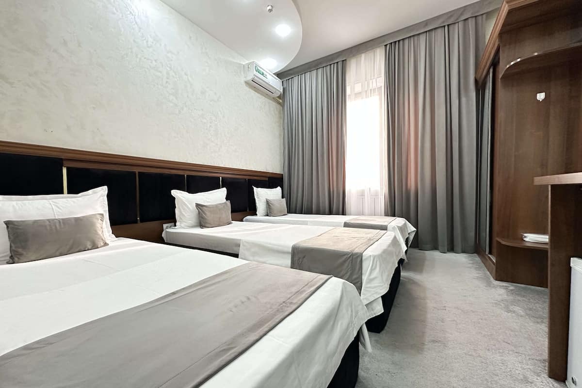 Une chambre d’hôtel avec trois lits, une tête de lit en bois, une grande fenêtre avec des rideaux et un appareil de climatisation fixé au mur.