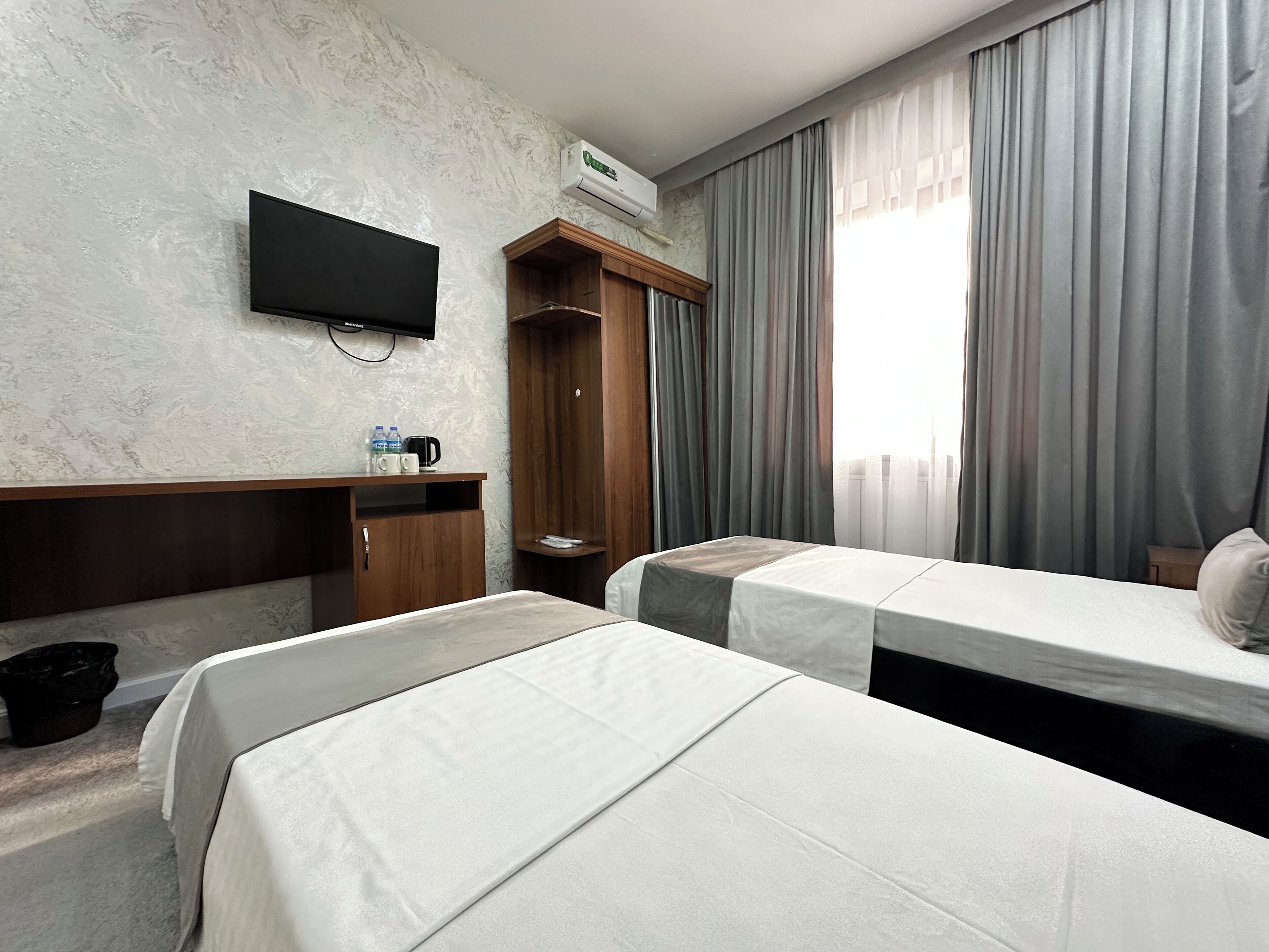 A hotel room with two beds, a TV, a wardrobe, a desk with a coffee maker, and a window with curtains.