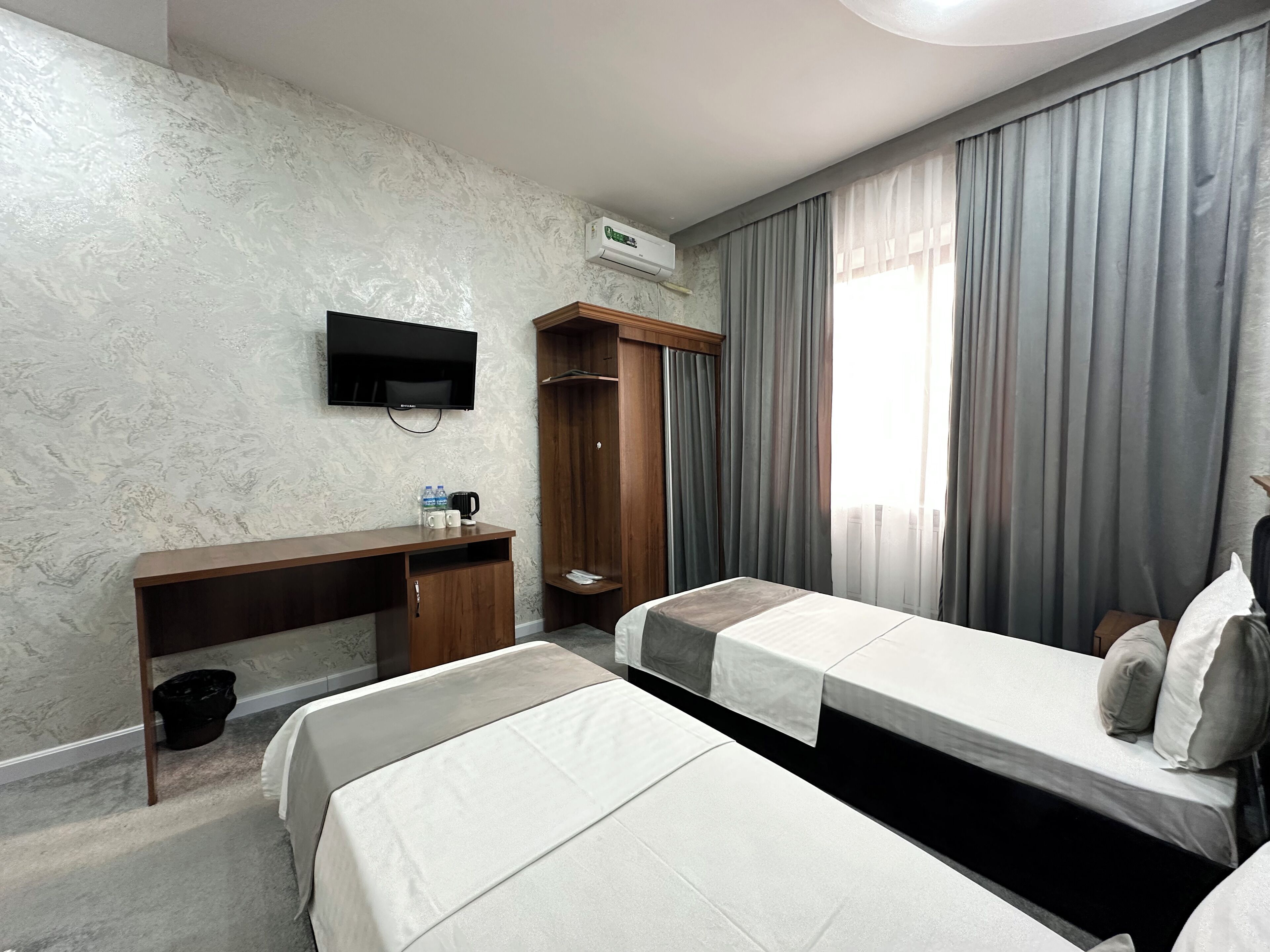 A hotel room with a marble-patterned wall, a flat-screen TV, a wooden desk with a chair, a wardrobe, a bed with a white mattress, and a window with grey curtains.