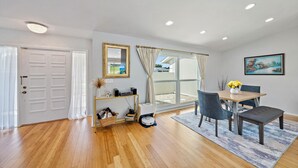 A modern living room with a wooden floor, a dining area with a table and chairs, a large window with curtains, and a painting on the wall.