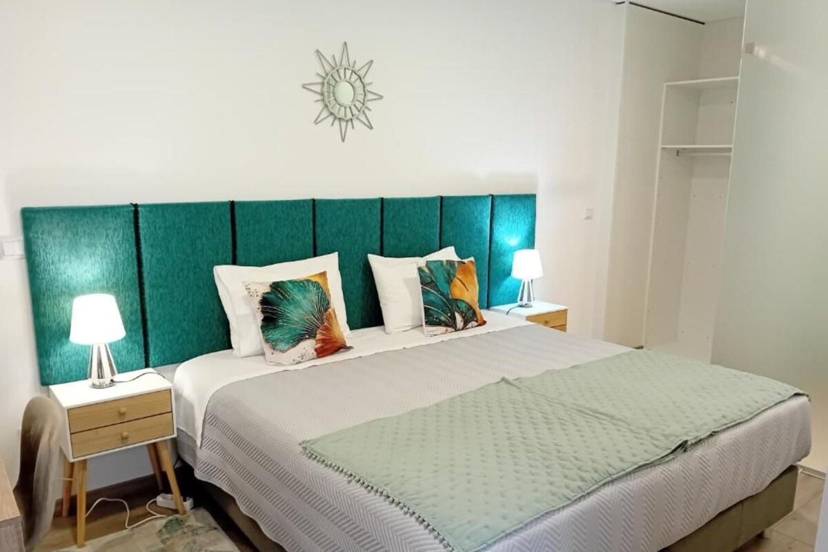 Un dormitorio con cabecera color turquesa, cama con colcha verde y blanca, dos lámparas de noche y una mesita de noche con lámpara.