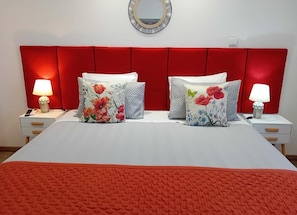 Una cama con cabecera roja, dos mesitas de noche con lámparas y almohadas estampadas con motivos florales.