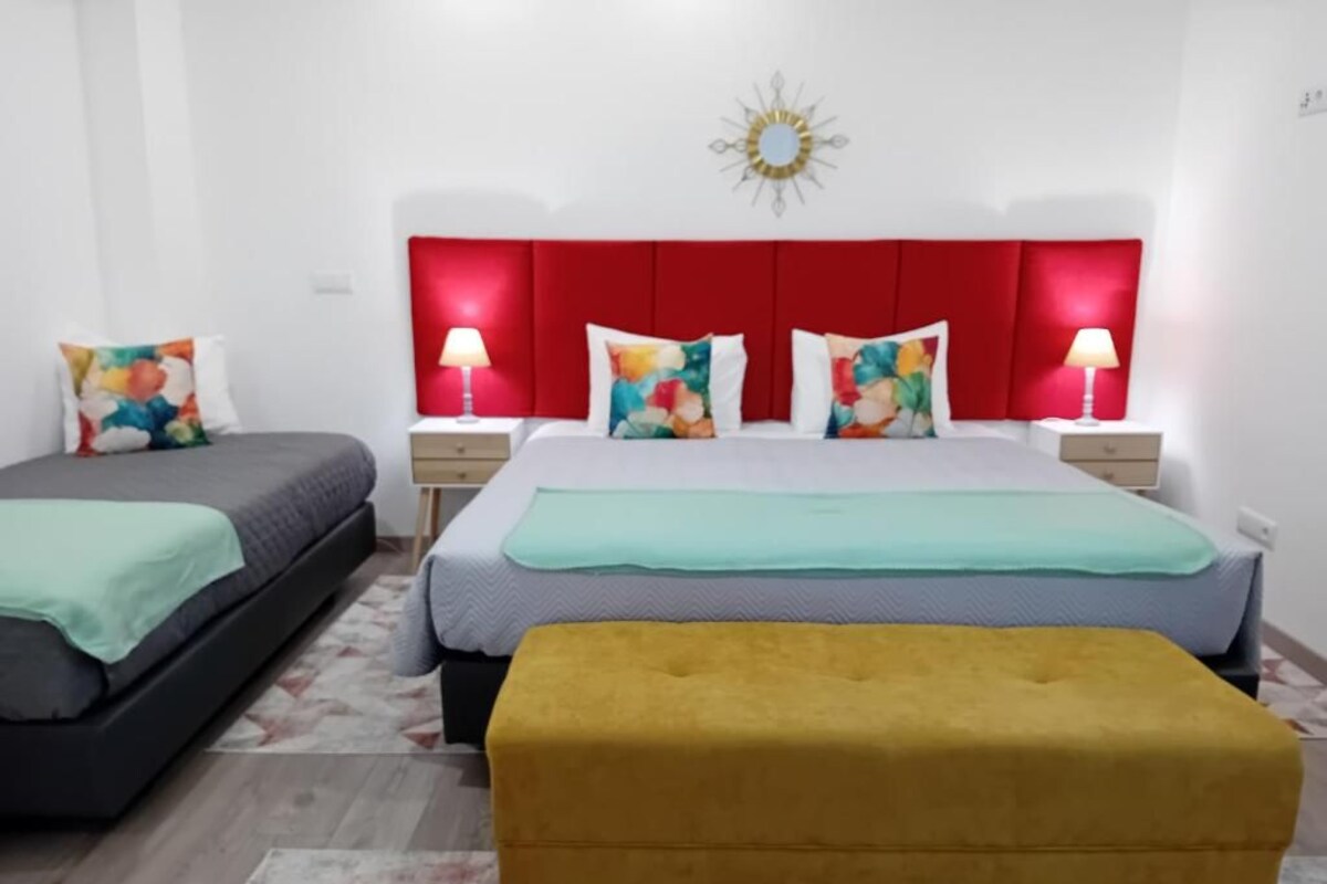 Una habitación de hotel con cabecera roja, dos camas, un reposapiés amarillo y un adorno de pared.