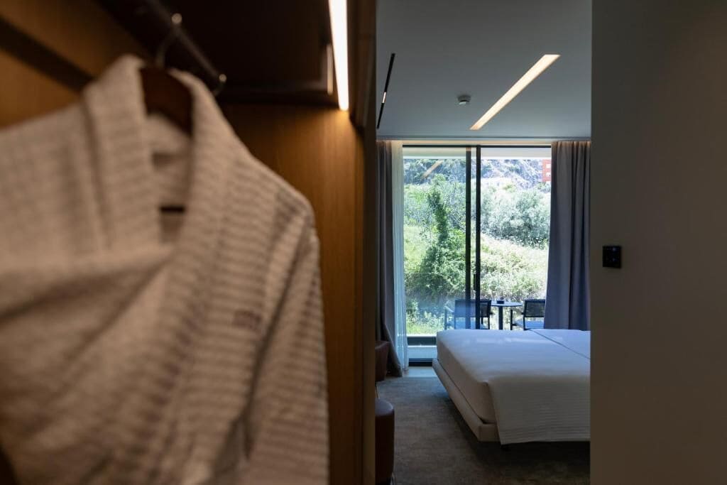 A hotel room with a bed, a view of the outdoors, and a robe hanging on a door.