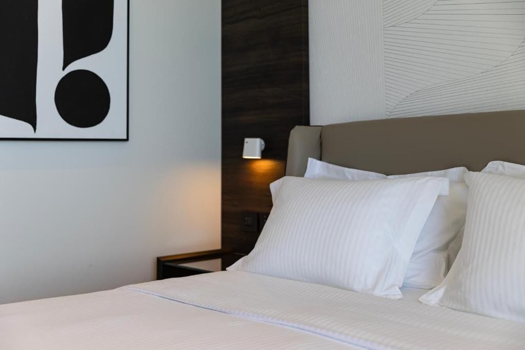 A neatly made bed with white linens, a dark headboard, and a wall-mounted light fixture.