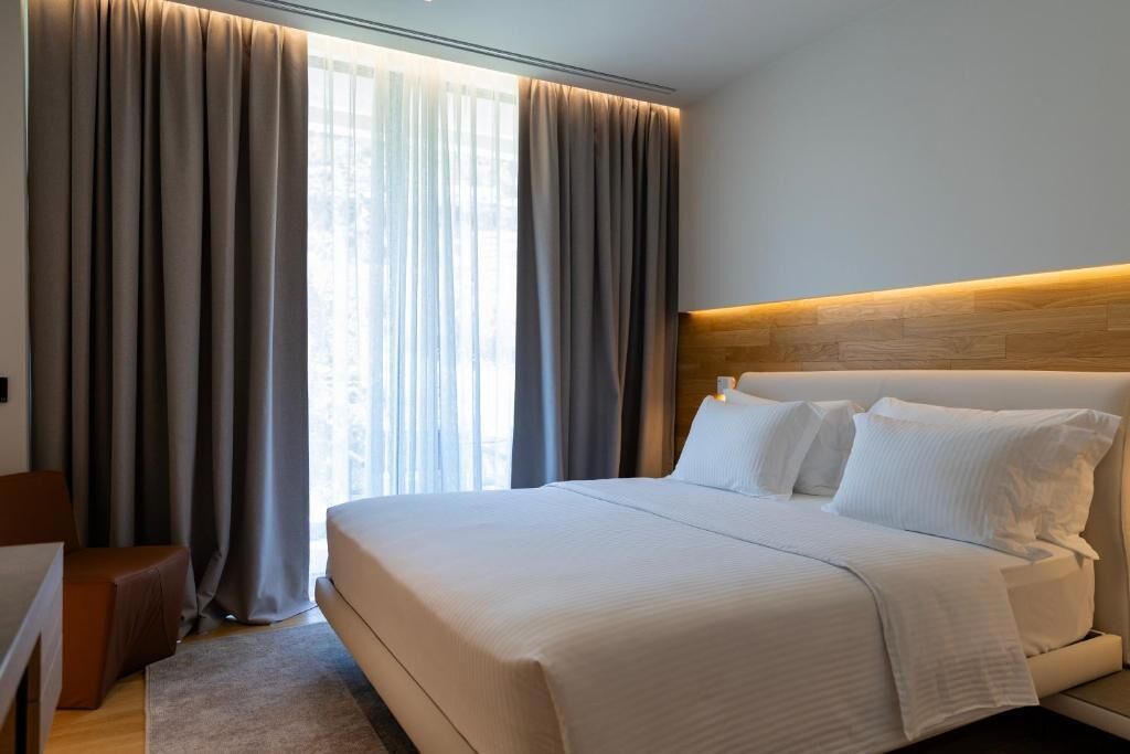 Una habitación de hotel con una cama grande, un cabecero de madera y vistas a través de cortinas translúcidas.