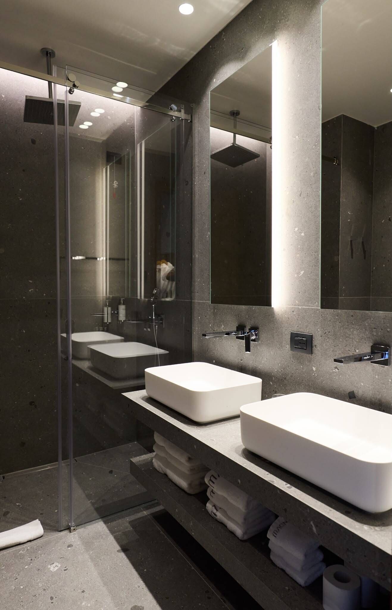 Un baño moderno con ducha envuelta en vidrio, dos lavamanos blancos y un espejo grande.