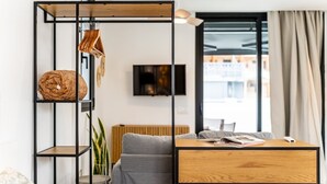 A modern living room with a wooden coffee table, grey sofa, and a black-framed shelving unit with a rolled-up rug and hanging clothes.