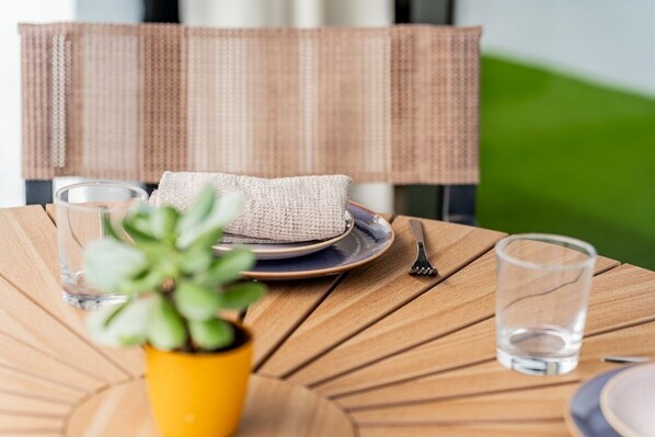 Mesa servida con platos, cubiertos y un vaso de agua, con una planta y el respaldo de una silla tejida al fondo.