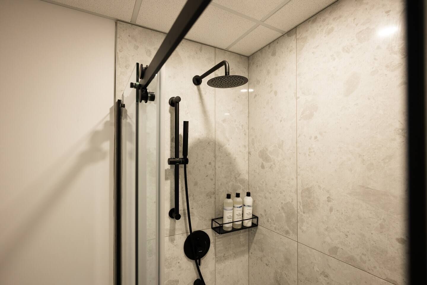 Ducha moderna con cabezal de ducha manual y otro de lluvia, puerta de cristal y repisa con artículos de aseo.