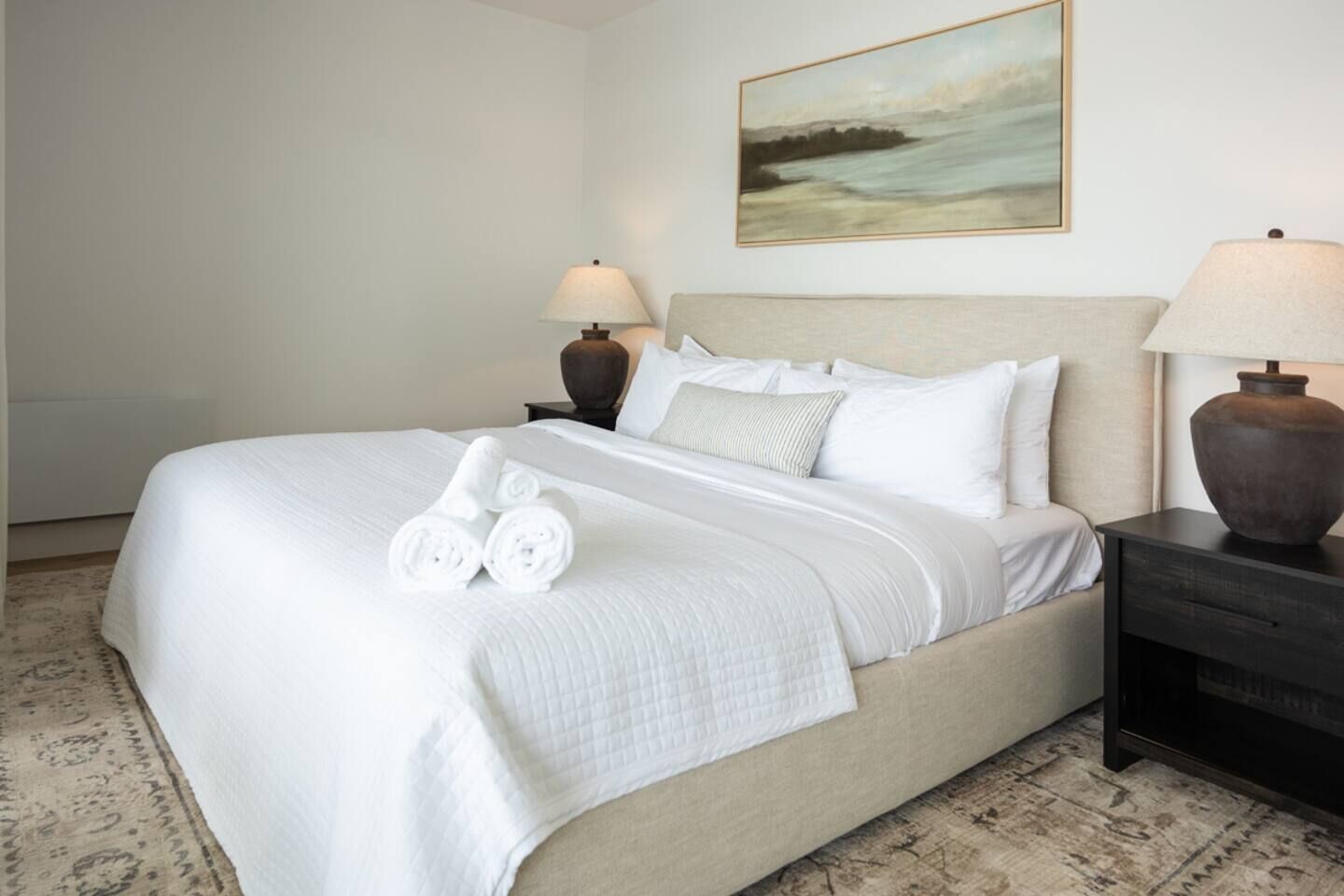 Cama bem arrumada com lençóis brancos e toalhas enroladas, cabeceira bege e um quadro de paisagem na parede.