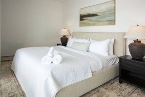 A neatly made bed with white linens and rolled towels, a beige headboard, and a painting of a landscape on the wall. - La Capitainerie - Vétérans 201 - Bord de l’eau (Lac-Mégantic)