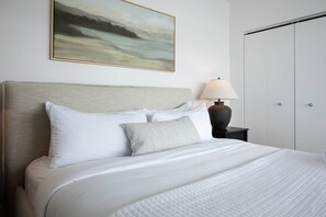 Een opgemaakt bed met witte beddengoed en een gestreept kussen, een bedlampje en een schilderij aan de muur.