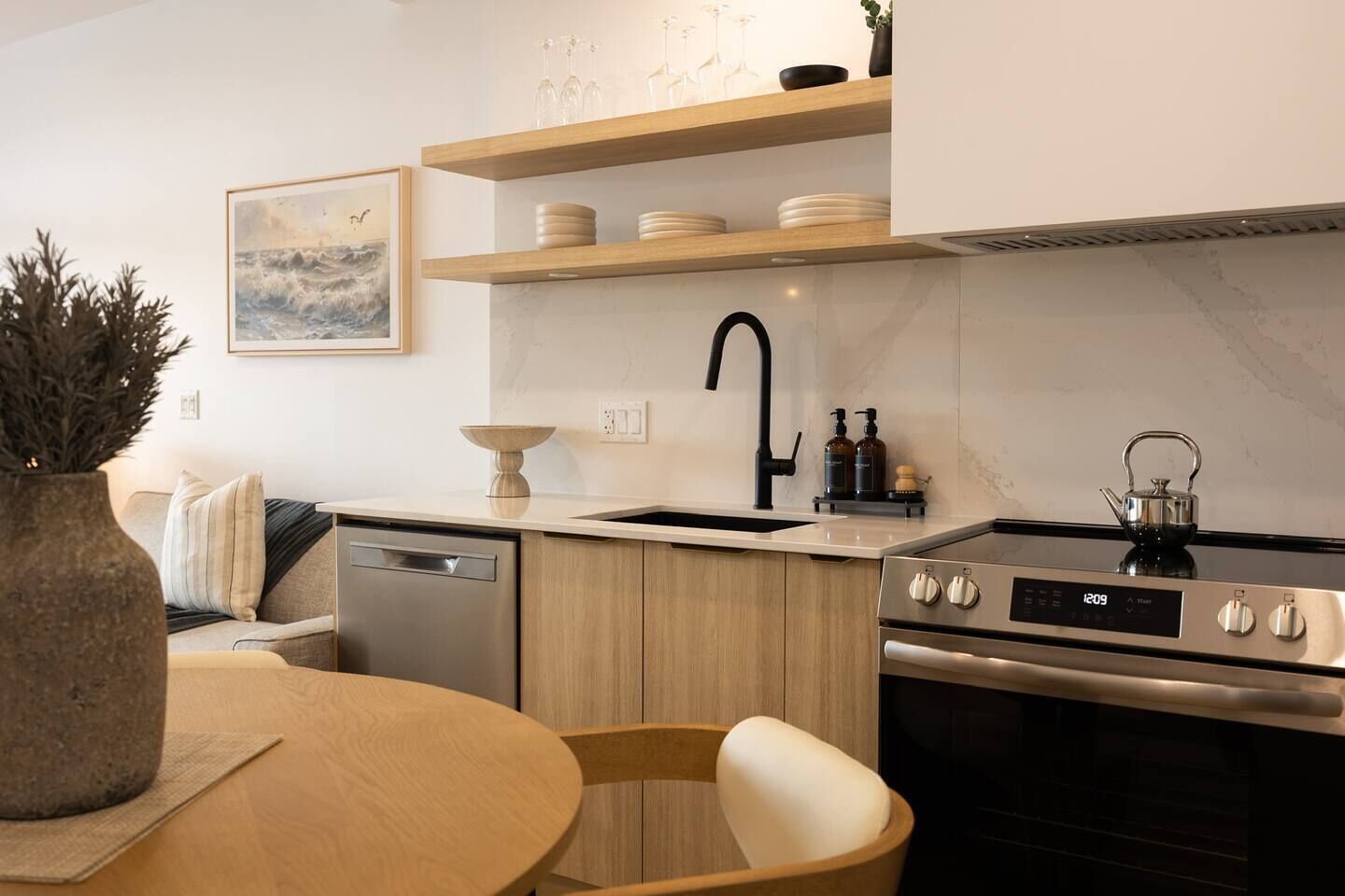 Een moderne keuken met een vaatwasser, fornuis en spoelbak. Er is een houten eettafel met stoelen en een vaas met planten.