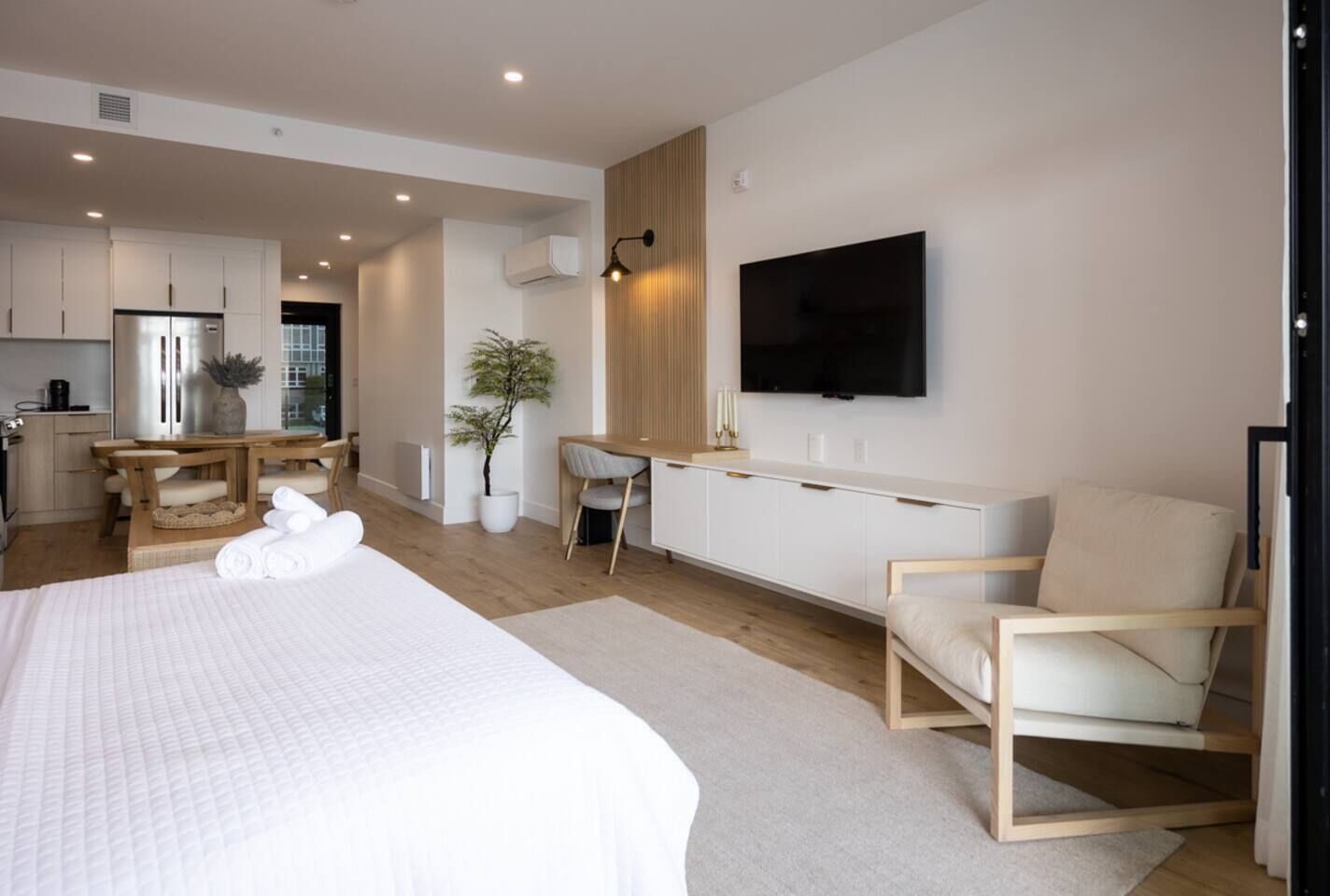 Una habitación de hotel moderna con una cama grande, un escritorio con una silla, un televisor montado en la pared y una kitchenette al fondo.