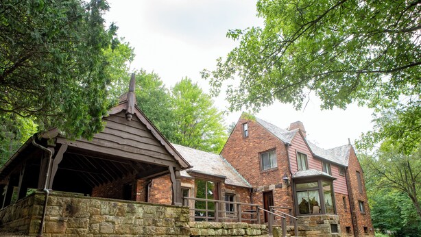 A rustic stone building with a sloped roof and wooden beams, surrounded by lush greenery.