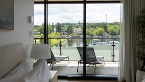 A room with a large window overlooking a river, a bed, a chair, and a table.