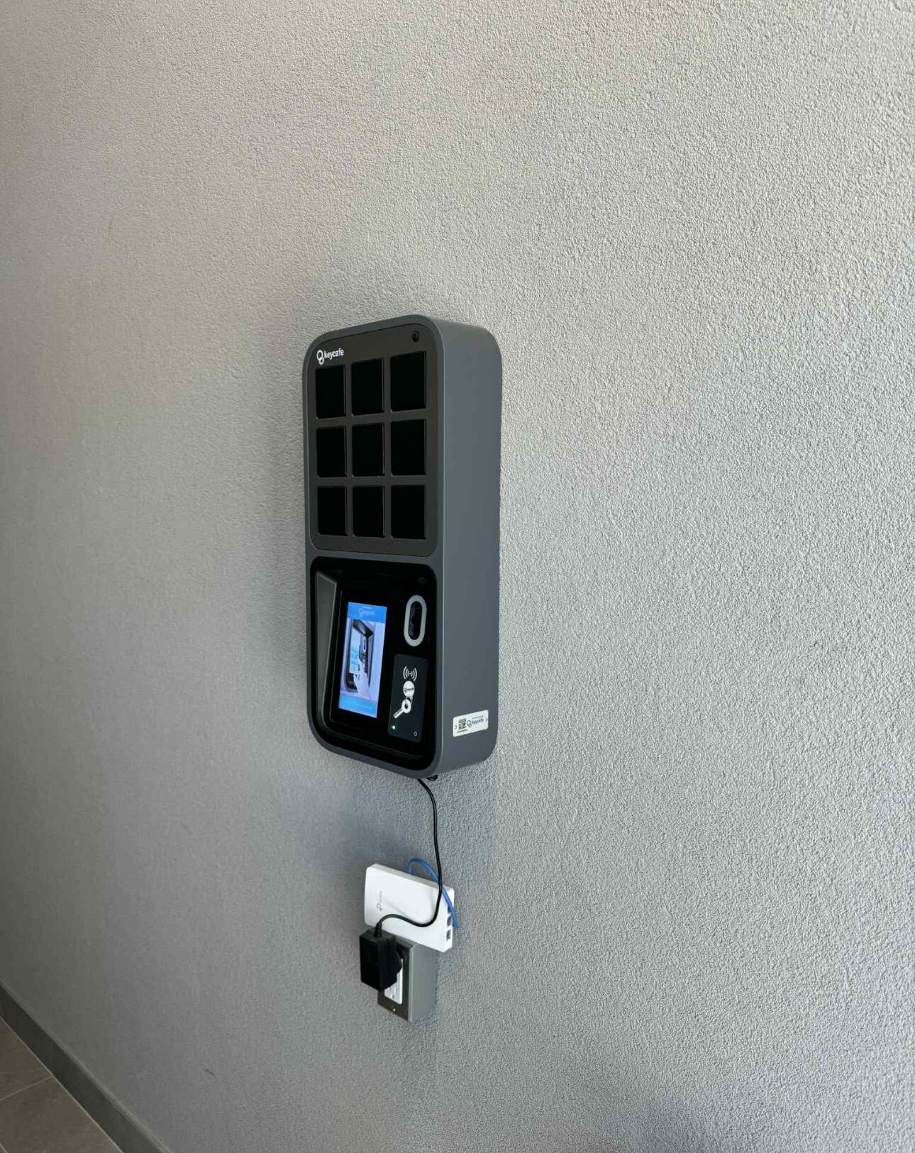 A wall-mounted security device with a keypad and a screen, connected to a power outlet.