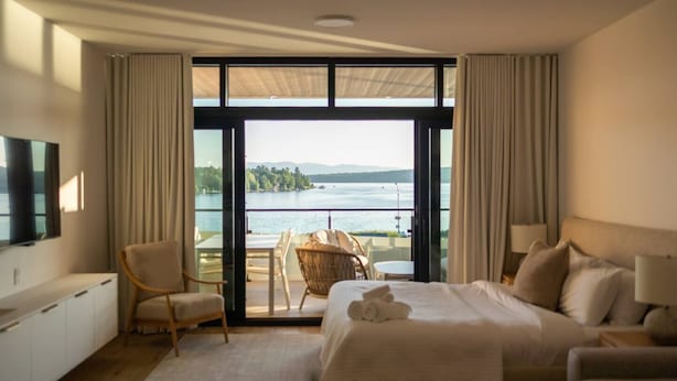 Una camera d'albergo con un grande letto, una televisione, un balcone con tavolo e sedie e vista su un lago.