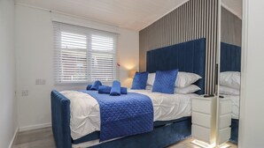 A modern bedroom with a large bed, a bedside table, a mirror, and a window with blinds. - Jackos Lodge (Windermere)