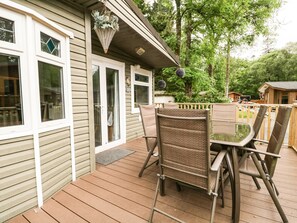 A deck with a table and chairs, a hanging basket, and a view of a wooded area. - Jackos Lodge (Windermere)