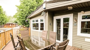 A deck with a glass-top table and wicker chairs, overlooking a wooden deck and a building with a number '9' sign. - Jackos Lodge (Windermere)