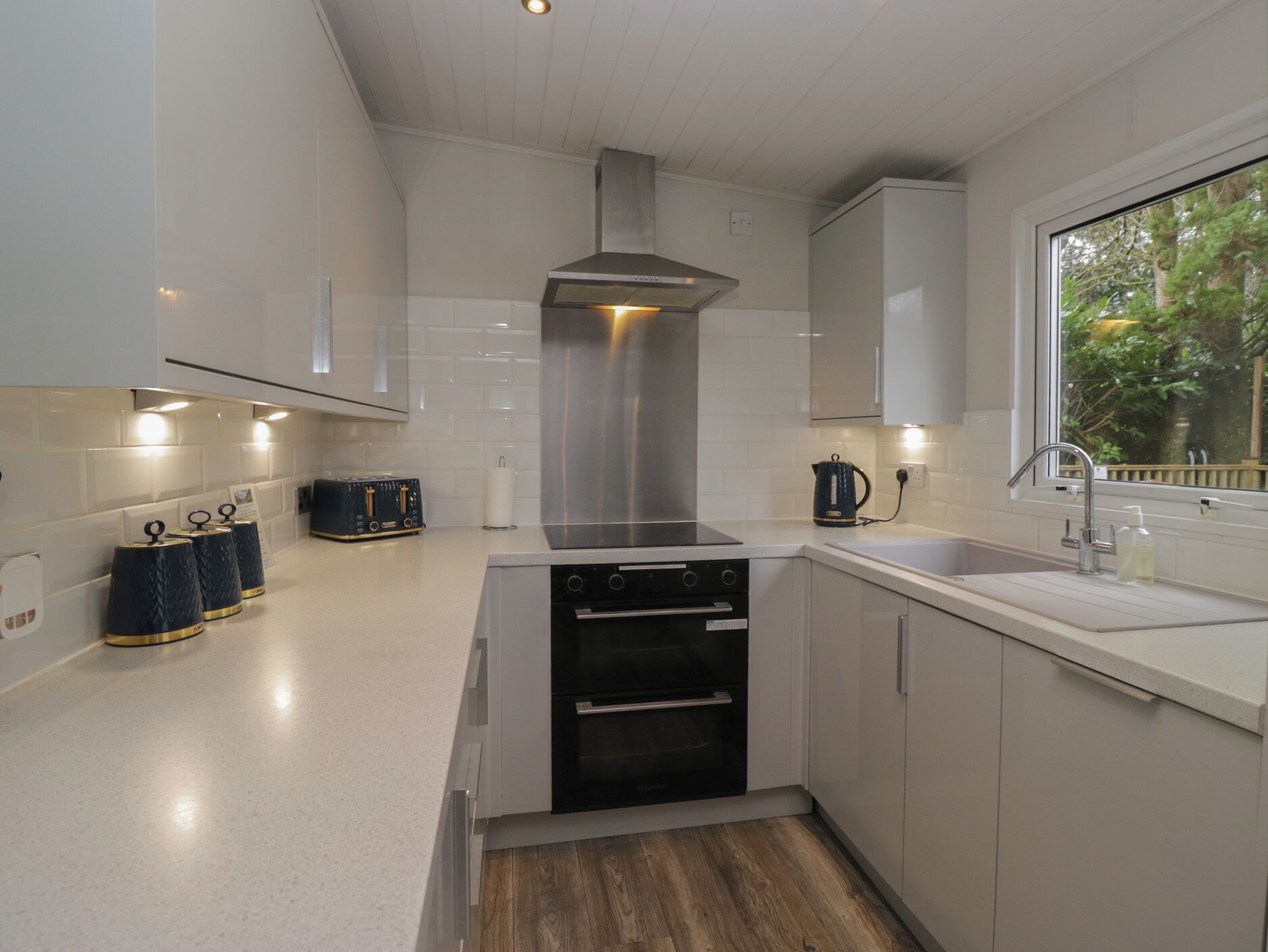 A modern kitchen with white cabinets, a stainless steel range hood, a built-in oven, and a window with a view of trees.